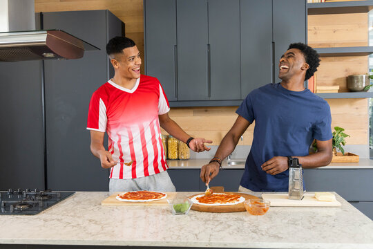 Cooking diverse male friends spreading sauce on pizza dough at home kitchen island, with utensils