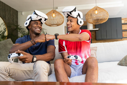 Fist-bumping male friends sitting on sofa in living room, with VR headsets and controllers