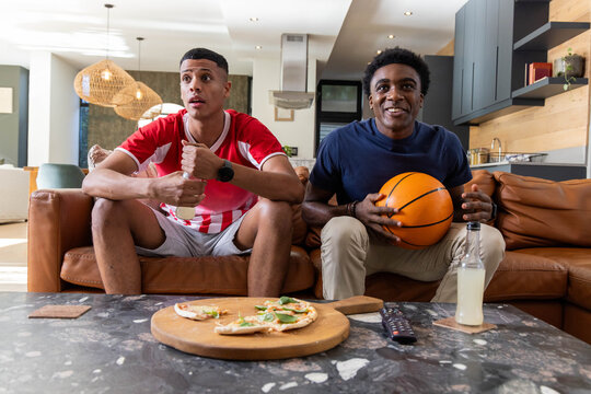 Sitting diverse male friends chatting on brown couch in living room, with basketball and pizza