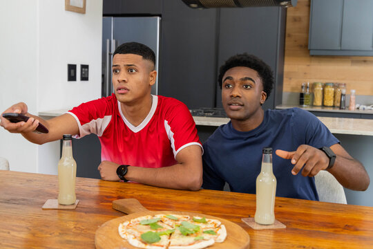 Diverse male friends holding remote and pointing at TV in home kitchen, with pizza and beverages