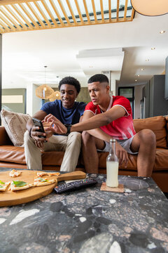 Sharing smartphone, diverse male friends sitting in modern living room, with bottle, pizza, remote