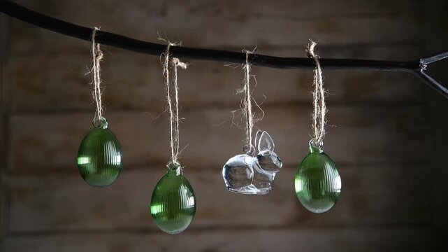 Glass Easter decorations transparent bunny and three ribbed green eggs are suspended on rustic-style twine from dark branch against a warm wooden background, creating atmosphere of cozy spring decor