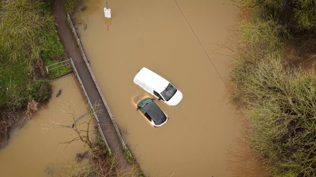 UK flooding submerging countryside road and trapping vehicles in muddy river water. Drone flying above inundated street near footbridge with cars sinking during heavy rain. Extreme weather hitting