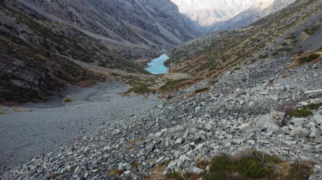 Low altitude drone flight over rugged rocks toward a turquoise alpine lake; a metaphor for resilience and overcoming