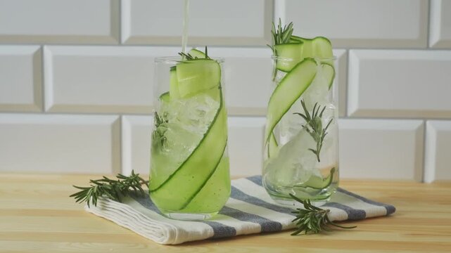 Cucumber rosemary mocktail pours into ice filled glass beside second garnished glass on striped napkin in bright kitchen.