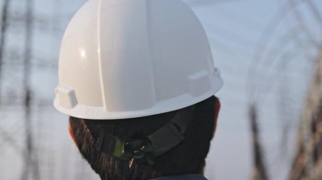 Power line utility worker safety helmet high visibility vest near electrical grid transmission tower and high voltage utility pole energy