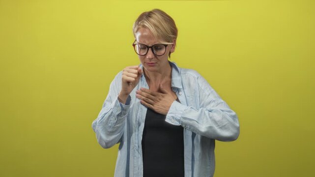 Blond woman wearing glasses and light blue shirt coughing into fist with hand on chest in studio against yellow backdrop; illness discomfort.