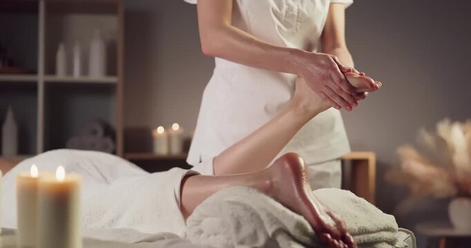 Massage foot woman masseur hands spa therapy. Foot reflexology with warm oil on towel as candles glow, easing stress in a serene spa setting indoors. Concept of calm healing spa relaxation.