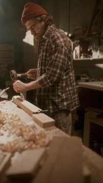 Bearded craftsman carving wooden plank with chisel and mallet in workshop, striking material on workbench during woodworking process. Vertical format footage