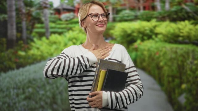 Woman holding books, hand on chest on a forest path wearing glasses and striped sweater; gratitude reflection.