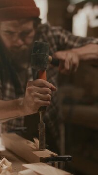 Bearded craftsman carving timber plank with chisel and mallet in carpentry workshop, focusing on details during woodworking process. Vertical clip, rack focus