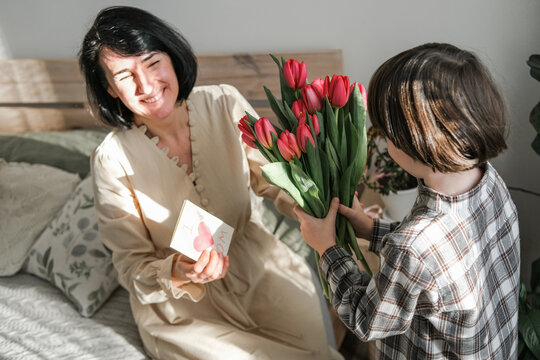Happy mother receiving a bouquet of red tulips and handmade greeting card from her young son. Emotional family moment during Mother's Day celebration in sunny cozy home interior