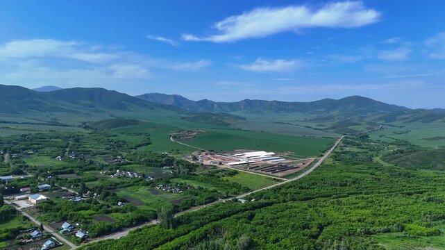 Drone footage captures a quaint small village with houses, roads, and paths nestled at the foothills of low mountains covered in green trees and meadows. Adjacent to the vast Bukhtarma Reservoir