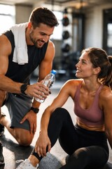 Obraz premium Male fitness trainer giving water to female client resting after workout in gym. Concept of fitness support, personal training session, recovery process, healthy lifestyle motivation.