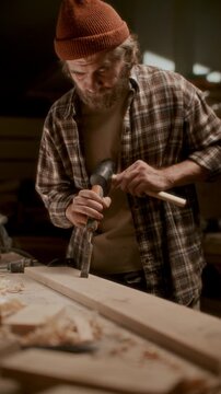 Skilled woodworker in plaid shirt and knit hat carving mortise into timber plank with hand chisel and mallet in carpentry workshop. Vertical format video