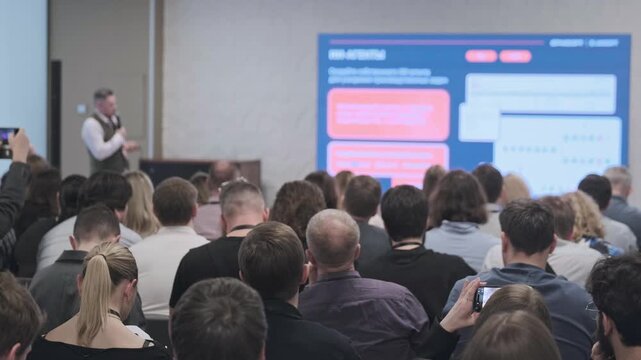 Business conference presentation with attentive audience watching speaker present technology slides on large screen, networking, learning, professional event atmosphere.
