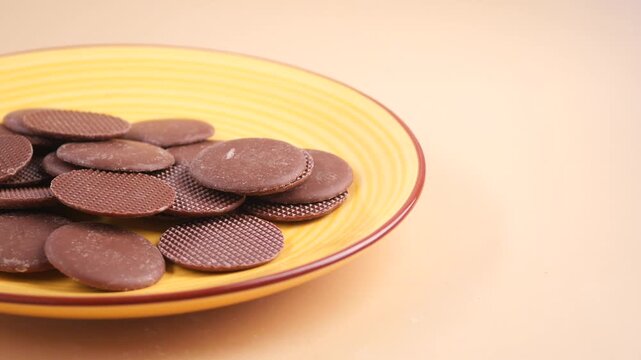 Dark chocolate coin chips rotating on yellow ceramic plate against beige background motion.