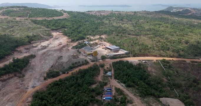 Forward aerial view revealing a concrete mixing plant with yellow silos and aggregate piles next to a hillside quarry, surrounded by lush tropical forest and unpaved red dirt roads.