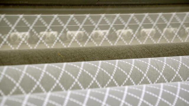 Close up of patterned fabric and textile rolls on a production line, geometric stitched material moving under industrial machine rollers in a factory for upholstery and home textile manufacturing
