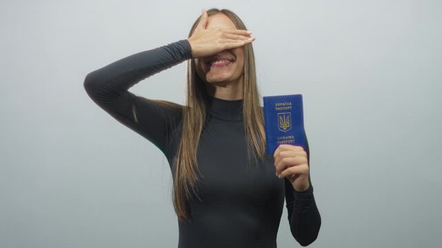 Woman holding ukrainian passport upright and covering her eyes with her hand in a studio with plain gray backdrop, passport emblem visible; shyness.