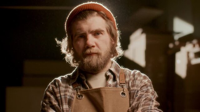 Professional woodworker in knit hat and apron posing for camera with confidence in sunlit carpentry workshop. Video portrait, chest-up shot
