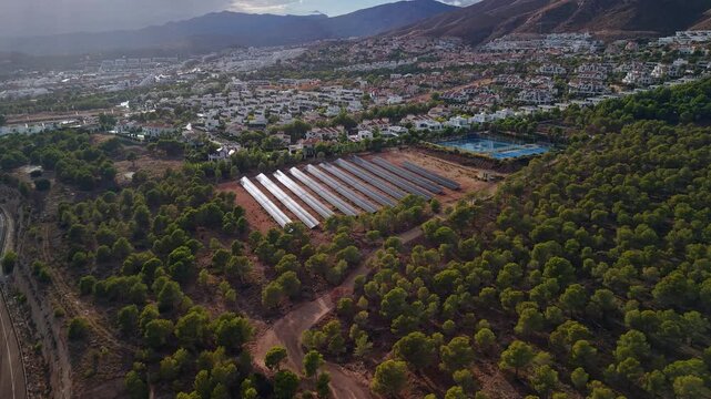Aerial view of solar power plant near residential area with rows of photovoltaic panels surrounded by forest and hills