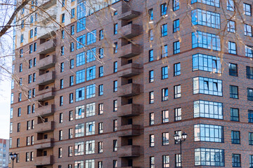 Fototapeta premium Modern apartment building with brick facade and glass windows. Balconies line each floor in repeating geometric pattern. Reflections of sky and trees shimmer on glass panes