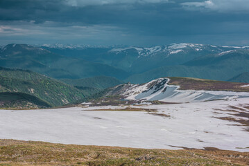 Sunlit landscape with small lake among snowfields, thawed ground, forest and snow cornice on long spur with view to big snowy mountain range far away under cloudy sky. Sunlight and shadows of clouds. © Daniil
