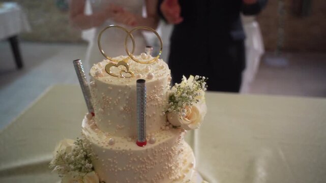 Elegant wedding cake with rings and white flowers, held by newlyweds in celebration
