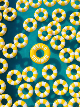 Hello Summer! Summer vacation, holiday, summertime, travel, tourism concept. A top-down view of yellow and white striped inflatable swim rings floating on a bright turquoise blue pool water surface.	