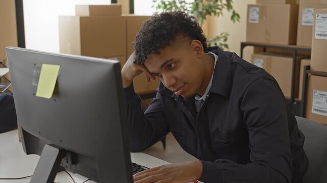 Man typing at computer among stacked parcels, resting head on hand in a packing area building; tired concentration.