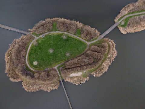 Aerial view of a small island with vibrant green grass, surrounded by a ring of brown vegetation and interconnected by pathways, standing out against the dark water, Overgooi, Flevoland, Netherlands.