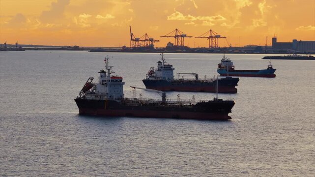 Aerial drone view of multiple oil tankers anchored near an industrial port at sunset with cranes and harbor infrastructure visible on the horizon, Limassol, Cyprus