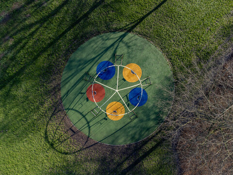 Aerial view of a vibrant, multi-colored merry-go-round spinning amidst the green grass, casting shadows from the trees, Messiaenplantsoen Speeltuin, Almere, Flevoland, Netherlands.