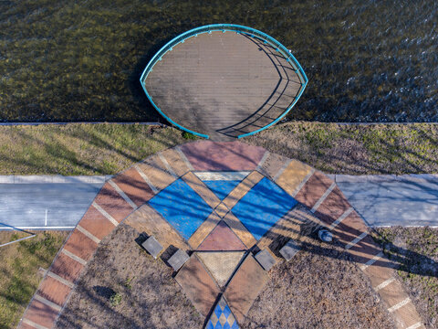 Aerial view of a decorative walkway with geometric patterns leading to an oval-shaped structure on the water, reflecting the sunlight, Maastrichtkwartier, Flevoland, Netherlands.