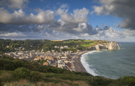 Aerial view of the dramatic cliffs meeting the sea, with a charming town nestled below under a sky streaked with clouds, Etretat, France.