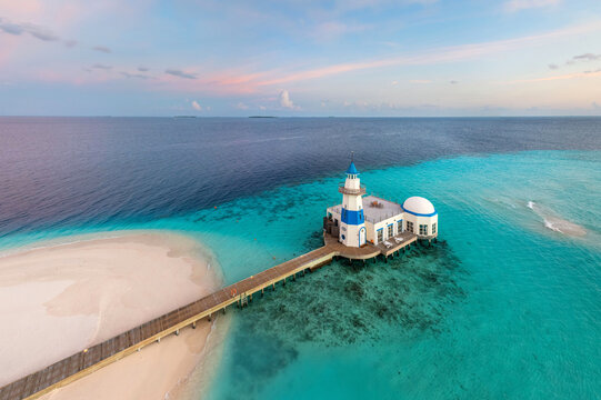Aerial view of a vibrant turquoise sea contrasting with the deep blue, surrounding a pristine white sandy beach and a striking white and blue mosque connected by a wooden pier, Maldives, Maldives.