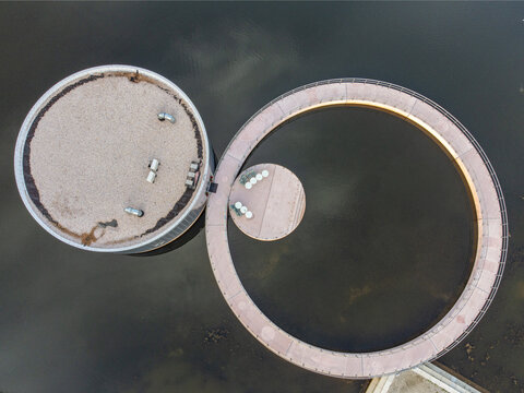 Aerial view of two circular structures connected by a winding path over dark waters, creating a serene contrast of shapes and textures, Utopia, Flevoland, Netherlands.