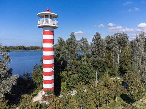 Aerial view of the striking red and white striped lighthouse standing tall against the lush greenery and calm waters, Utopia, Flevoland, Netherlands.