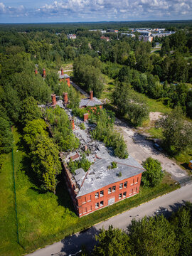 Aerial view of a red brick building with broken windows and overgrown vegetation, surrounded by lush green trees, creating a striking contrast between decay and nature, Liepaja, Latvia.