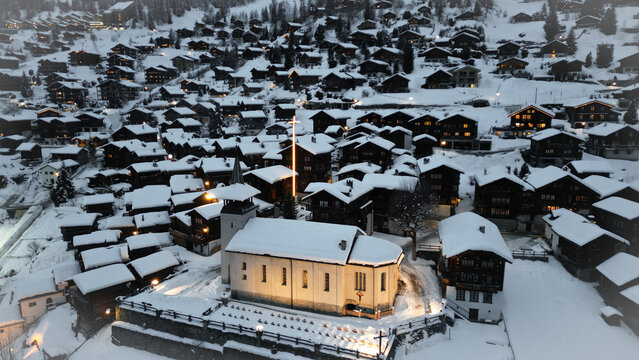 Aerial view of a snow-covered village with a brightly lit church standing prominently amidst the dark, rustic chalets, Grimentz, Valais, Switzerland.