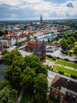 Aerial view of the Holsten Gate framed by vibrant green trees, with the old town's red rooftops and the twin spires of Lubeck Cathedral in the distance, Lubeck, Schleswig-Holstein, Germany.