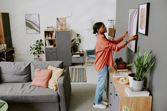 Young Black woman arranging framed artwork on wall in living room, styling home interior for decor update and organization concept. Wide shot, suitable for home decor marketing and lifestyle branding