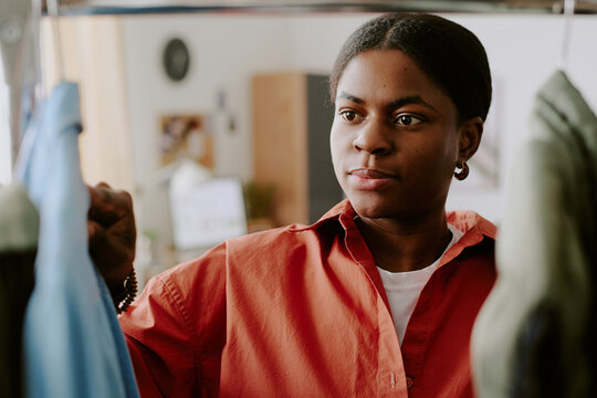 Medium shot of young Black woman selecting clothing on rack, comparing options for purchase decision. Supporting retail marketing, personal styling, consumer choice, ecommerce campaigns