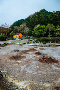 View of steaming fumaroles dotting the volcanic landscape contrasting with the vibrant yellow house nestled against the lush green hillside, Furnas, Azores, Portugal.