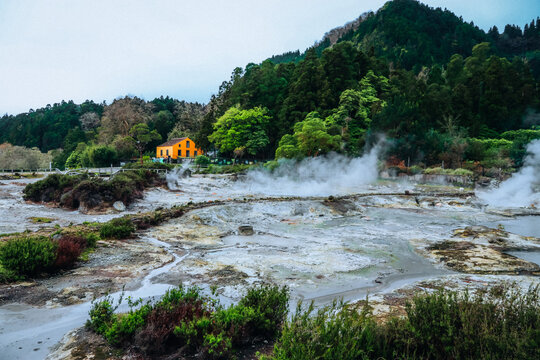 View of steaming vents rising from the ground amidst lush greenery, with a bright yellow house perched on a hillside in Furnas, Azores, Portugal.