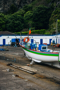 View of a vibrant fishing boat with green and red trim sits on the concrete, with white buildings and lush greenery rising behind, Caloura, Acores, Portugal.