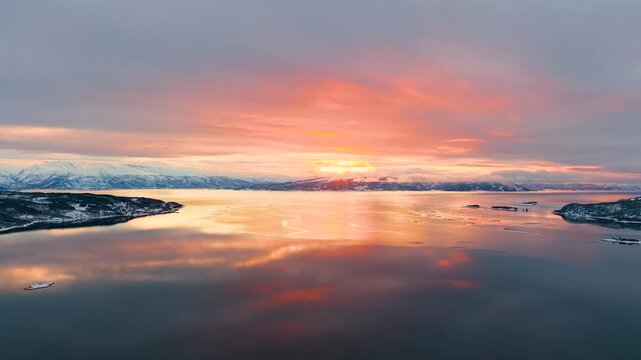 Breathtaking aerial view of a vibrant sunset reflecting on the calm waters of a Norwegian fjord, surrounded by snowcapped mountains in Lofoten.
