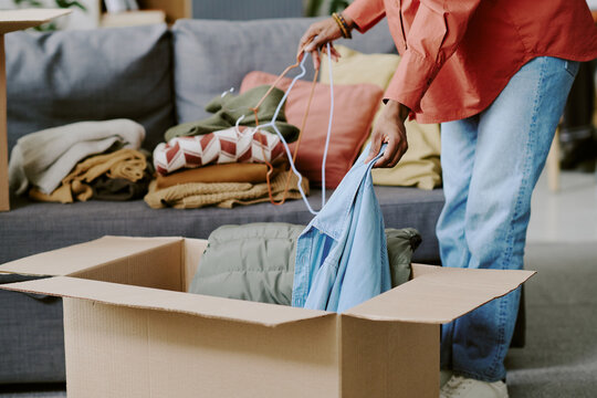 Black woman packing folded clothes into cardboard box while sorting hangers at home, preparing donation or relocation. Useful for charity, decluttering, moving, resale campaigns