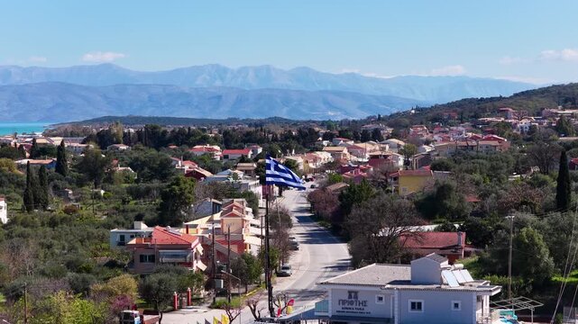 Aerial view of Acharavi village in Corfu, Greece. Large Greek flag waving over town street with the Ionian sea, coastal landscape, and majestic mountains in the background on a sunny day.